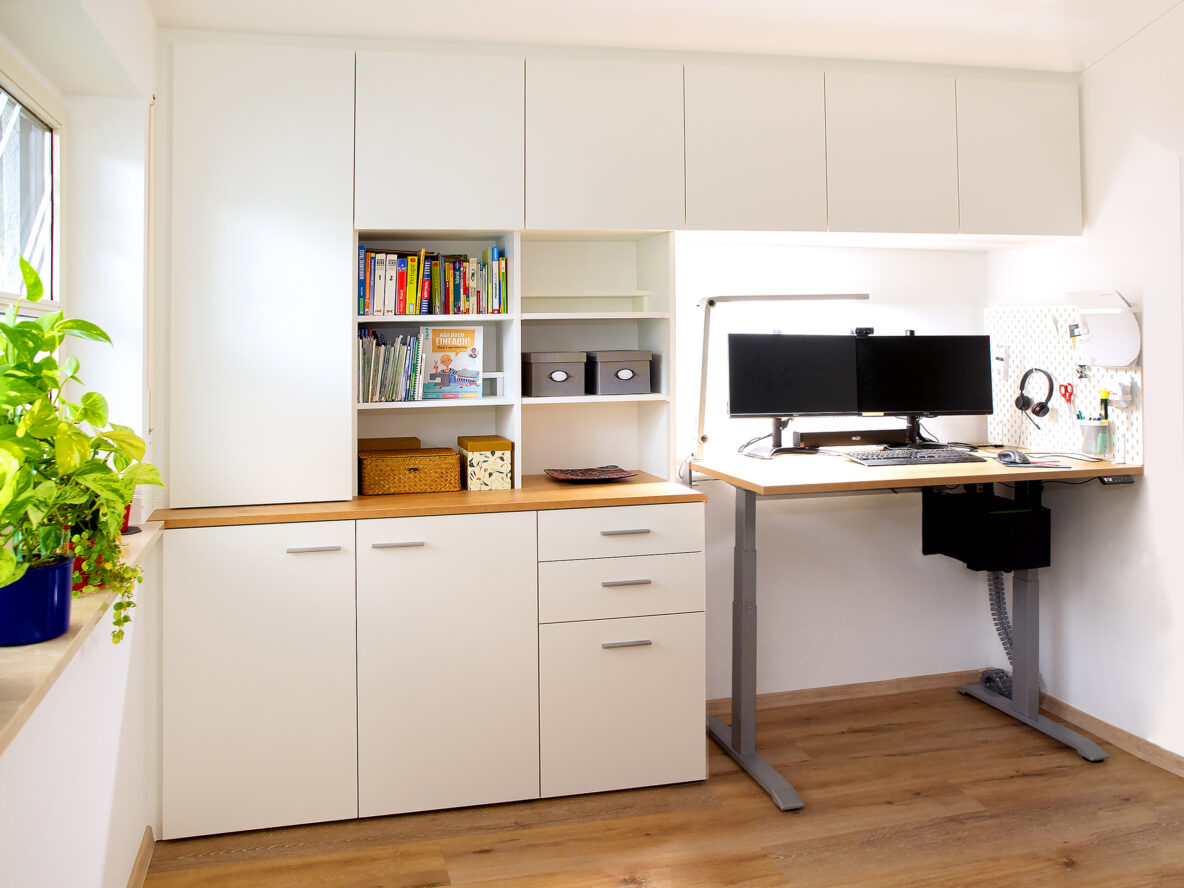 Homeoffice mit einem höhenverstellbaren Schreibtisch, mit einem Tischüberbau, mit Schränken und mit weiß beschichteten Regalen. Bild wurde mit KI retuschiert.