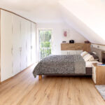 Einrichtung für Schlafzimmer in der Dachschräge mit einem grauen Doppelbett und einem weißen Einbauschrank. Bild wurde von KI retuschiert.