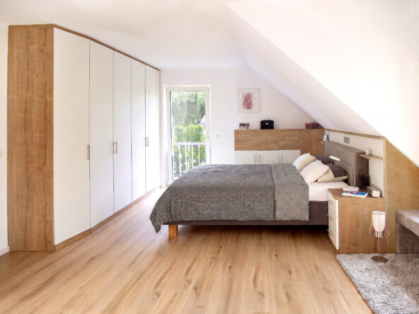 Einrichtung für Schlafzimmer in der Dachschräge mit einem grauen Doppelbett und einem weißen Einbauschrank. Bild wurde von KI retuschiert.